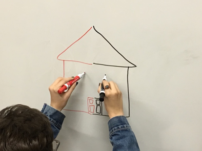 Student is drawing on a whiteboard using both hands. The image the student is drawing is a house. The student is holding a black marker in their right hand and a red marker in their left hand