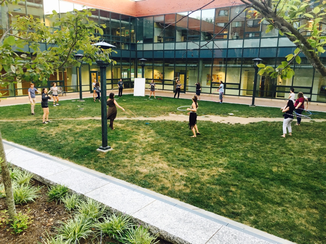 Students are standing in a circle and playing with hula hoops on a grassy courtyard.